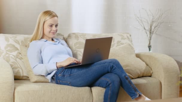 Schöne blonde Frau sitzt auf einer Couch mit Notizbuch auf dem Schoß, sie tippt aktiv darauf. ihr Zimmer ist voller Licht. — Stockvideo