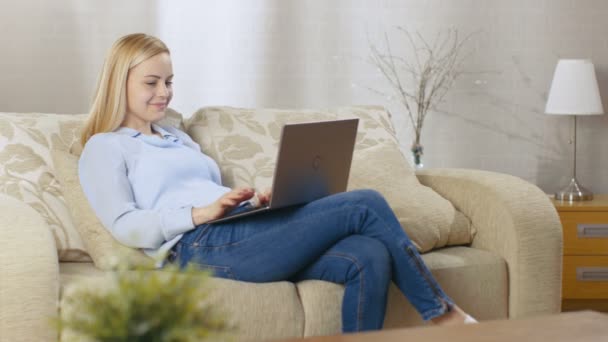 Piękna Blondynka kobieta siedzi na kanapie z notebooka na kolanach, ona aktywnie wpisuje na nim. Jej pokój jest pełen światła. — Wideo stockowe
