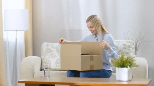 Beautiful Young Blonde at Home Opening Cardboard Box while Sitting on Her Sofa. — Stok Video