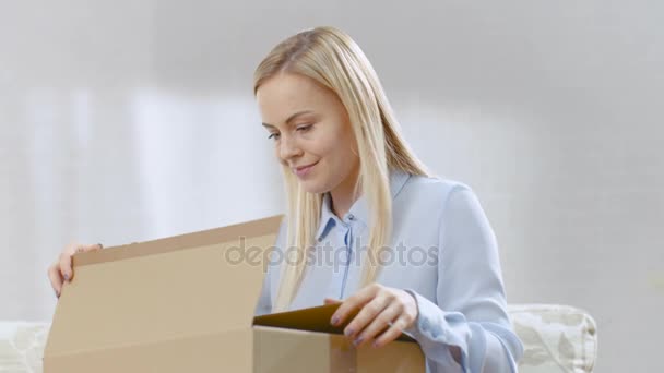 Beautiful Young Woman at Home abre una caja de cartón mientras está sentada en un sofá en su luminosa sala de estar. Ella sonríe . — Vídeo de stock