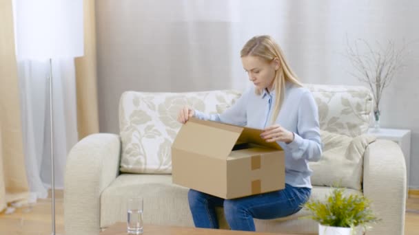 Beautiful Young Woman at Home abre una caja de cartón mientras está sentada en un sofá en su luminosa sala de estar. Ella sonríe . — Vídeo de stock