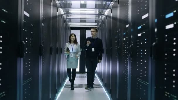 Kaukaski kobieta mężczyzna i Azjatycka to technicy spaceru w centrum danych z wierszy do stojaka, serwery. Mają one dyskusji, ona trzyma komputer typu Tablet. — Wideo stockowe