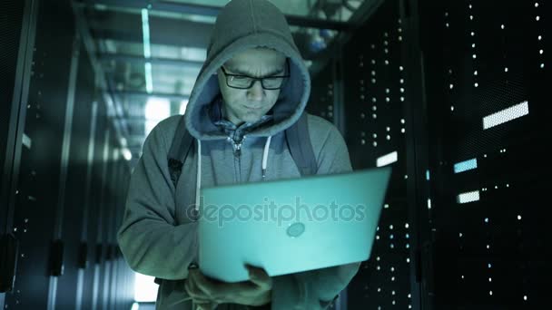 Hacker em um capuz em pé no meio do data center cheio de servidores de rack e Hacking-lo com seu laptop . — Vídeo de Stock