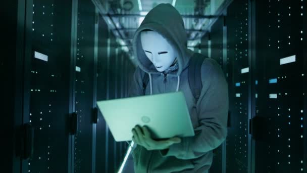 Крупный план Хакер в маске в капюшоне, стоящий в центре обработки данных, полный стеллажных серверов и взломать его с помощью ноутбука . — стоковое видео