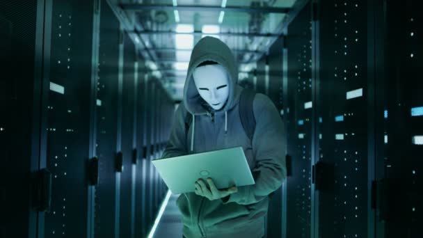 Mid Shot of a Masked Hacker in a Hoodie Standing in the Middle of Data Center full of Rack Servers and Hacking it with His Laptop. — Stock Video