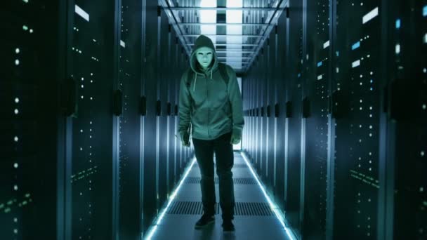 Lång skott av en maskerad Hacker i en Hoodie gående mot kameran i datacenter med rader av rackservrar av hans sidor. — Stockvideo