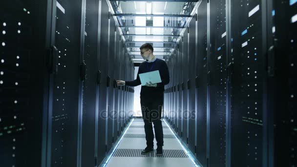 Vue frontale de l'ingénieur informatique marchant à travers le centre de données plein de serveurs de rack de travail. Il ouvre le cabinet de serveurs et se connecte à lui avec son ordinateur portable .