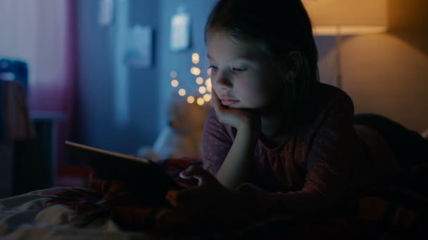 Sevimli küçük kız onun odasına gece, Tablet bilgisayar ile bir yatakta yatıyor. Açık onun gece lambası. — Stok video