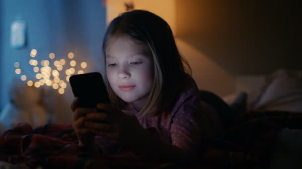 Мила маленька дівчинка в її кімнаті вночі, брехня на ліжку використовує смартфон. Її нічна лампа ввімкнулася . — стокове відео