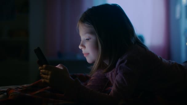 Το χαριτωμένο μικρό κορίτσι στο δωμάτιό της το βράδυ, βρίσκεται σε ένα κρεβάτι χρήσεις Smartphone. Τη νύχτα φανό ενεργοποιημένη. — Αρχείο Βίντεο