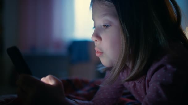 Cute Little Girl in Her Room at Night, Lies on a Bed Uses Smartphone (dalam bahasa Inggris). Layar Mengiluminasi Wajahnya . — Stok Video