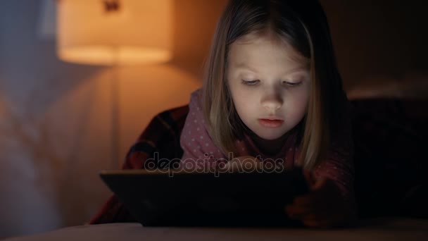 Söt liten flicka ligger i sin säng på natten, hon håller tablett dator. Hennes nattlampa är vände på. — Stockvideo
