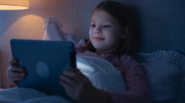 Mutlu küçük kız gece yatağında yatıyor ve Tablet bilgisayarda bir şey izliyor. Onun yatak ışığı yanıyor. 