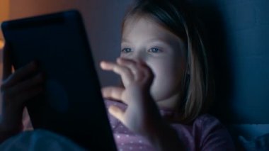 Sevimli küçük kız gece yatağında Tablet bilgisayar ile etkileşim kurar..