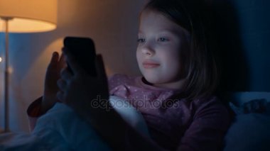 Sevimli genç kız yatak odasında yatıyor. O Smartphone kullanır. Onun gece ışığı yanar. O mutlu bir şekilde gülümsüyor.