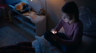 Sevimli küçük kız gece onun odasına. Smartphone kullanırken yatağında oturuyor. Onun yatak ışığı yanıyor.