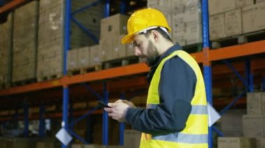 Depo işçisi kask giyiyor kullanır Smartphone ve depolama rafları satırları arasında yürüyor.