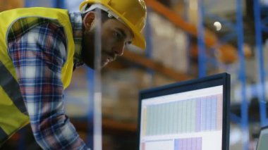 Baret takmış erkek Müfettiş elektronik tablolarında kişisel bilgisayarında doldurur. Palet raflar satır içeren büyük bir depo içinde.