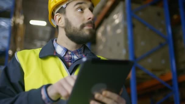 El topógrafo que usa casco duro sostiene la computadora de la tableta y cuenta la mercancía en el almacén llena de bastidores con cajas en ellos . — Vídeos de Stock