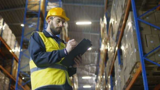 Supervisor que usa casco duro con portapapeles Rellena formularios en un almacén. Camina a través de filas de bastidores de almacenamiento con mercancía . — Vídeos de Stock