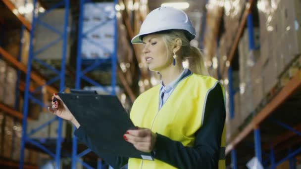 Il regolatore femminile che porta il cappello duro e che tiene il portablocco conta la merce che si leva in piedi in grande magazzino con i rack del pallet in esso . — Video Stock