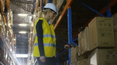 Kadın denetleyicisi kask giyiyor ve inceden inceye gözden geçirmek ile barkod tarayıcı mal. O büyük bir depo olduğunu palet raflar dolu.