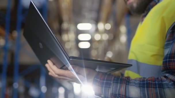 Warehouse Worker usa laptop. Ele está no meio de um grande centro de distribuição com grandes rachas de armazenamento e paletes neles. . — Vídeo de Stock