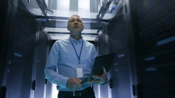 No data center, o engenheiro do servidor percorre o data center com linhas de rack de servidor enquanto mantém o laptop . — Vídeo de Stock
