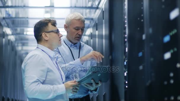 Dos técnicos de TI de pie en el centro de datos. Usan computadora portátil y tableta mientras están al lado del gabinete de rack de servidor abierto . — Vídeos de Stock