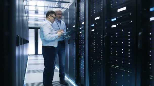 Two IT Engineers Standing in Working Data Center. They Use Laptop and Tablet Computer while Standing Beside Open Server Rack Cabinet. — Stock Video