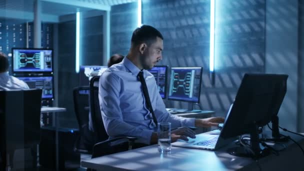 Primer plano del Ingeniero Técnico trabajando en su computadora con múltiples pantallas en la sala de monitoreo. En el fondo, sus colegas con gráficos y varios datos sobre sus monitores . — Vídeos de Stock