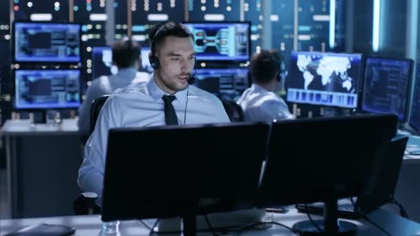 I System Control Center teknisk support Specialist taler i headset mens sidder ved sit skrivebord før flere skærme. Hans kollegaer arbejder i baggrunden i et rum fyldt med skærme . – Stock-video