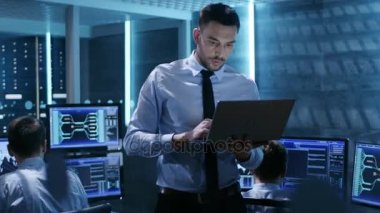 BT profesyonel Holding Laptop monitör oda yürür. Arka plan uzmanı meşgul iş istasyonlarında.