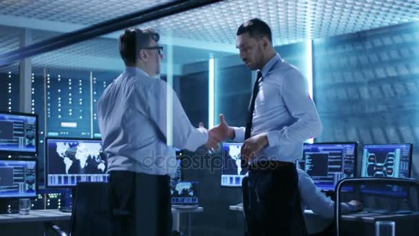 Deux ingénieurs en TI discutent et se serrent la main au milieu de la salle de surveillance remplie d'écrans de travail .