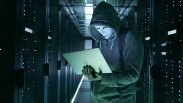 Primer plano de un hacker enmascarado con capucha parado en medio de un centro de datos lleno de servidores en rack y hackeándolo con su cuaderno . — Vídeos de Stock