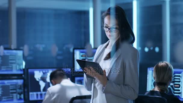 Молодая женщина-инженер использует планшет в центре системного управления. На заднем плане ее коллеги находятся на своих рабочих местах со многими дисплеями, показывающими ценные данные . — стоковое видео