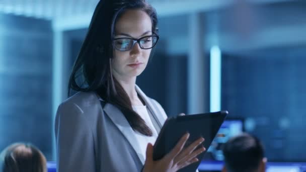 Jonge vrouwelijke regering werknemer bril gebruikt Tablet in System Control Center. Op de achtergrond zijn haar collega's op hun werkruimten met vele Displays toont waardevolle gegevens. — Stockvideo