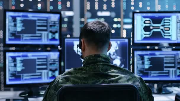 Back View of Military Teknisk support Professionel giver instruktioner ved hjælp af headset. Han arbejder i overvågningsrummet fuld af skærme, der viser forskellige oplysninger . – Stock-video
