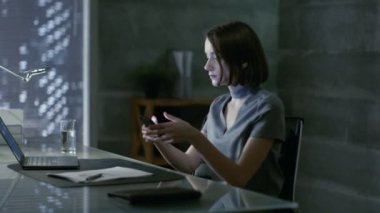 Başarılı genç kadın özel ofisinde masasında oturur ve Smartphone kullanır. Onun ofisi Minimalist tarzda yapılır, oldukça karanlık ve pencere vardır büyük şehir manzaralı.