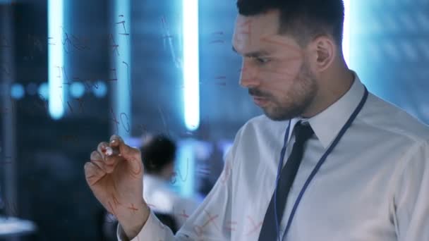 In der Systemzentrale schreibt der Chefingenieur mit rotem Marker auf eine Glastafel. im Hintergrund Überwachungsraum voller Arbeitsdisplays, die Daten und Personen zeigen, die arbeiten. — Stockvideo