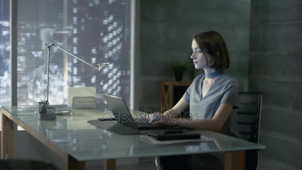 Jovem mulher bem sucedida senta-se em um escritório privado em sua mesa e funciona em um laptop. Seu escritório é feito em estilo minimalista, é bastante escuro e tem janela com vista para a cidade grande . — Vídeo de Stock