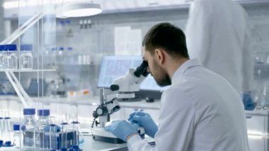 Modern laboratuvar araştırma bilim adamı ayarlar örnek bir Petri kabına cerrahi kerpeten ve mikroskop altında üstünde o görünüyor içinde. Meslektaşı gelir ve masadaki yerini alır.