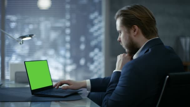Молодой и перспективный бизнесмен сидит за своим столом и типы на ноутбуке с зеленым макетом экрана. Ее офис выглядит современным с темными оттенками, большой город за окном . — стоковое видео