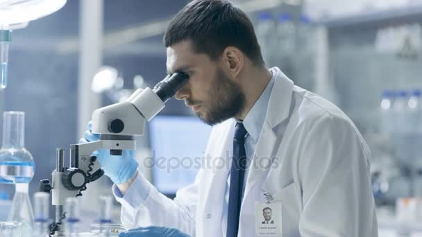 Forskning forskare ser ut i mikroskopet och skriver ner sina iakttagelser. Han arbetar i moderna laboratorier. — Stockvideo