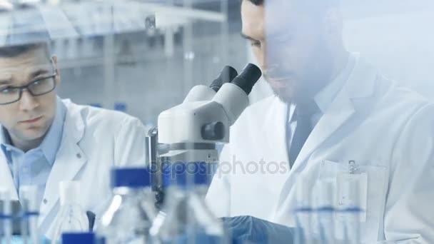 Modern bir laboratuarda iki bilim adamları deneyler. Baş araştırma bilim asistanı için mikroskop içinde gördüğü sonuçları belirler. — Stok video
