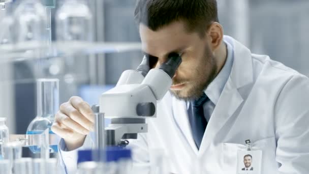 Research Scientist Adjusts His Microscope (en inglés). Está trabajando en un laboratorio moderno de gama alta con vasos de precipitados, cristalería, microscopio y monitores de trabajo que lo rodean . — Vídeo de stock