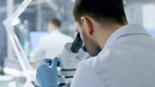 Sobre o ombro Vista de um cientista de pesquisa Olhando para o microscópio. Realiza experiências com os seus colegas de laboratório moderno. . — Vídeo de Stock