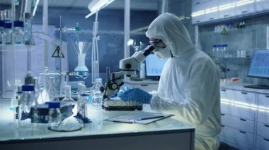 Güvenli yüksek düzeyinde araştırma laboratuvarı bilim adamı bu durumda bir Coverall örnekleri bir petri kerpeten ile ayarlar ve sonra onları mikroskop altında inceler.
