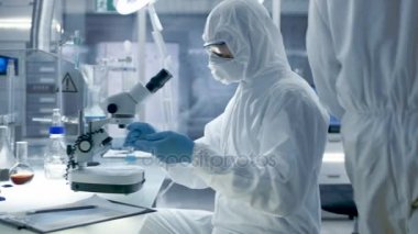Güvenli yüksek düzey laboratuvar bilim adamları bir Coverall içinde yapılan bir araştırma. Biyolog örnekleri bir petri kerpeten ile ayarlar ve sonra onları mikroskop altında inceler ve meslektaşı bir bilgisayarda sonuçları analiz eder..