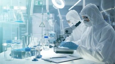 Güvenli yüksek düzey laboratuvar bilim adamları bir Coverall içinde yapılan bir araştırma. Kimyager bir petri kerpeten ile örnekleri ayarlar ve sonra onları mikroskop altında inceler ve meslektaşı sonuçları yazıyor.